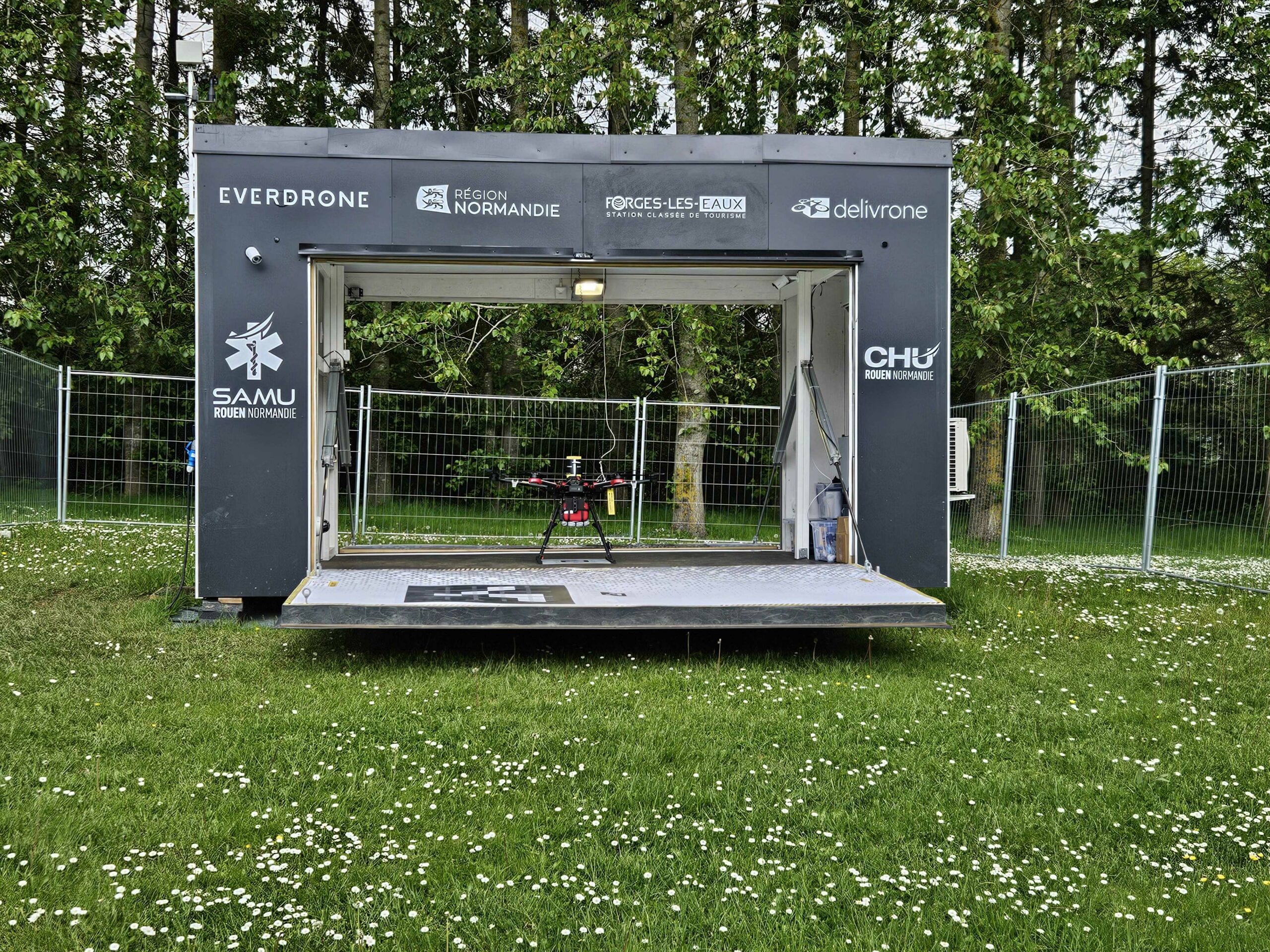 A drone hangar with logos for Everdrone, Région Normandie, Forges-les-Eaux, Delivrone, and CHU Rouen Normandie stands in a grassy area surrounded by trees. Inside the building, a drone is visible on the platform, ready for deployment.