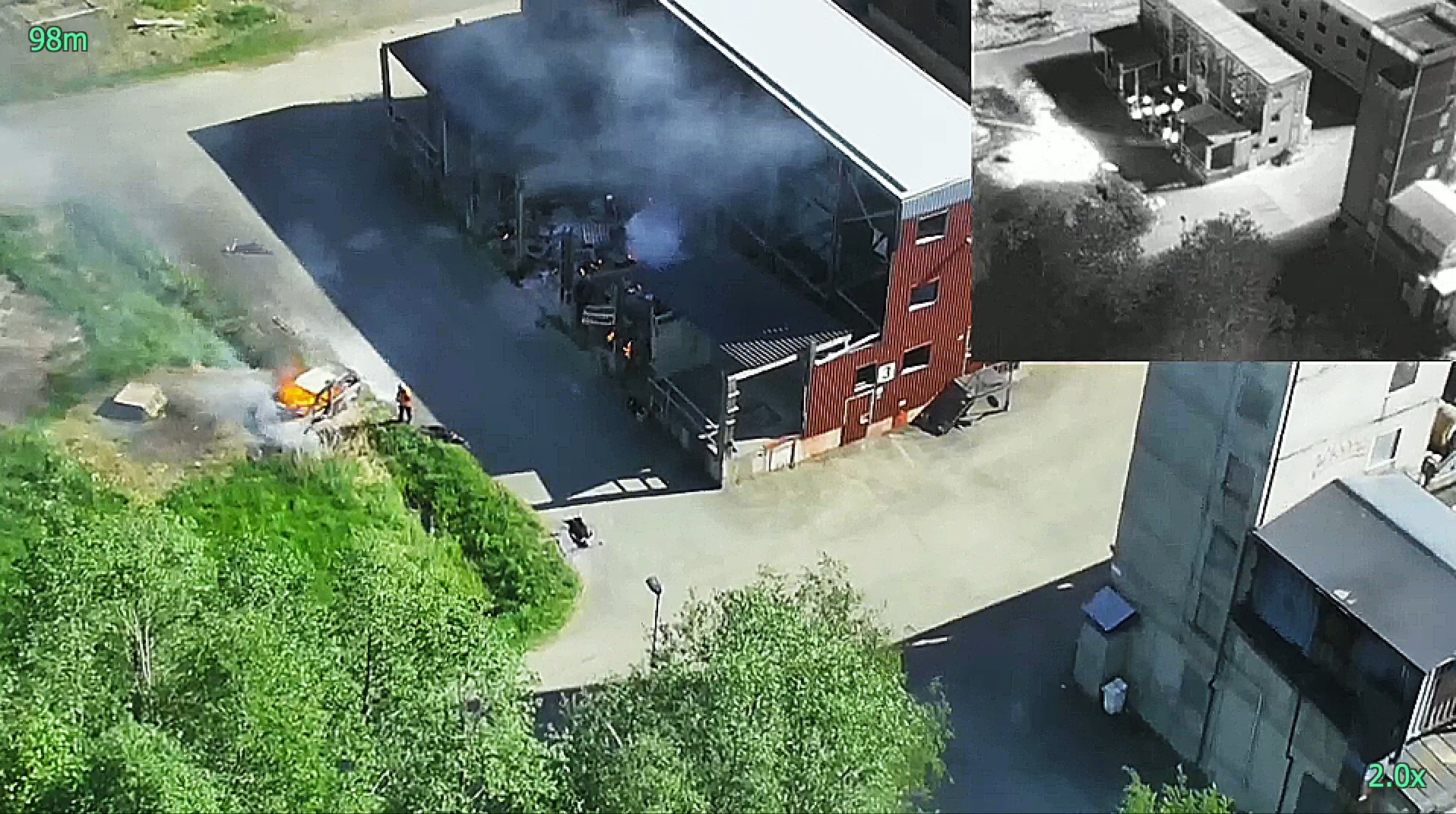 Aerial view of a fire response scenario captured by a drone. The main image shows a car on fire near a building with smoke billowing into the air, while firefighters work on the scene. An inset image in the top right corner displays a thermal view of the area, highlighting heat sources in the environment, providing a clear contrast between hot and cooler areas. The image demonstrates the use of drone technology for live monitoring and situational awareness in emergency response operations