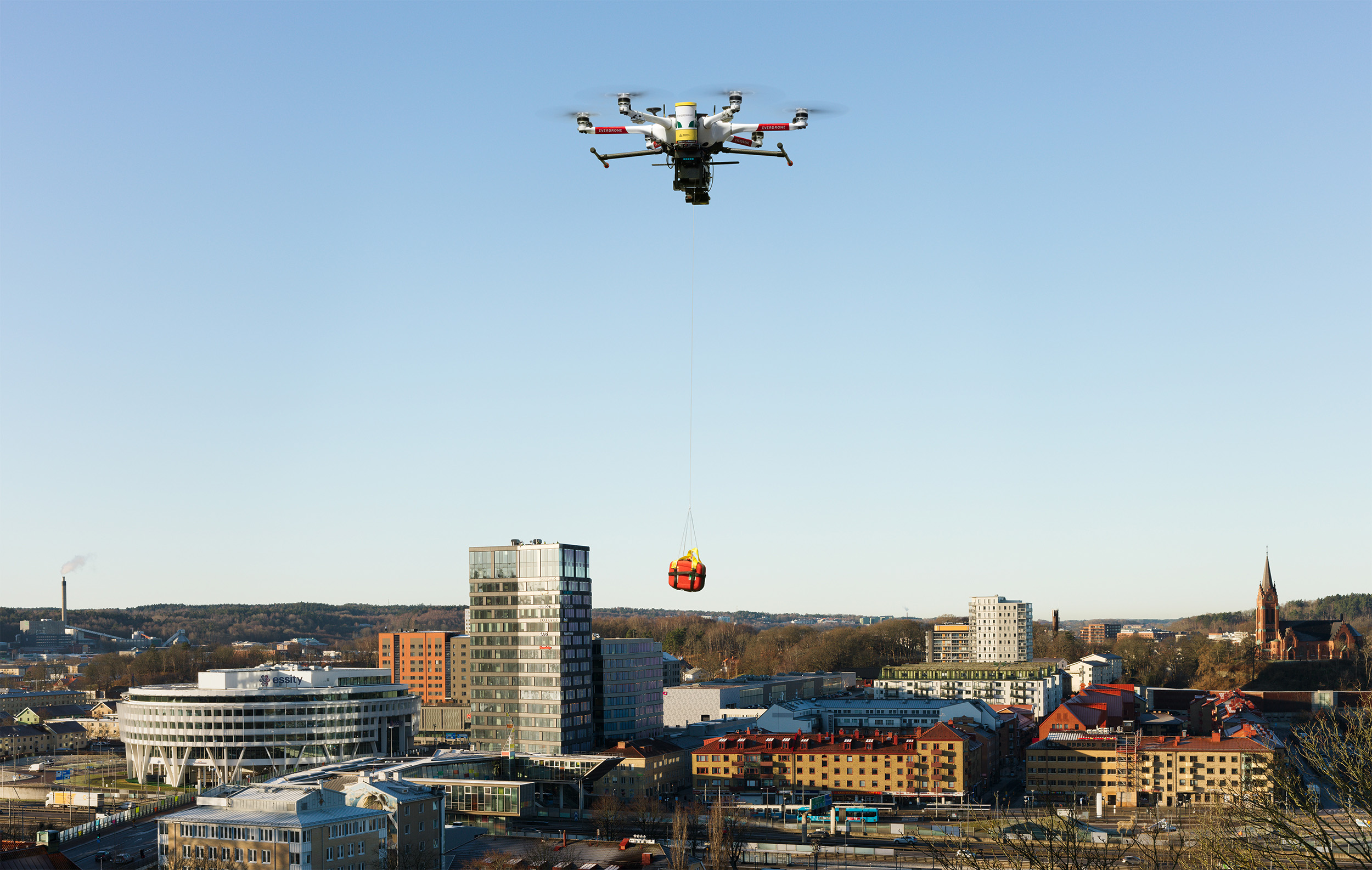 A drone hovers in the clear blue sky over a cityscape, carrying a red and yellow emergency medical package suspended by a cable. Below, modern office buildings, residential apartments, and a church with a tall spire are visible. The scene is bathed in warm sunlight, with long shadows suggesting early morning.