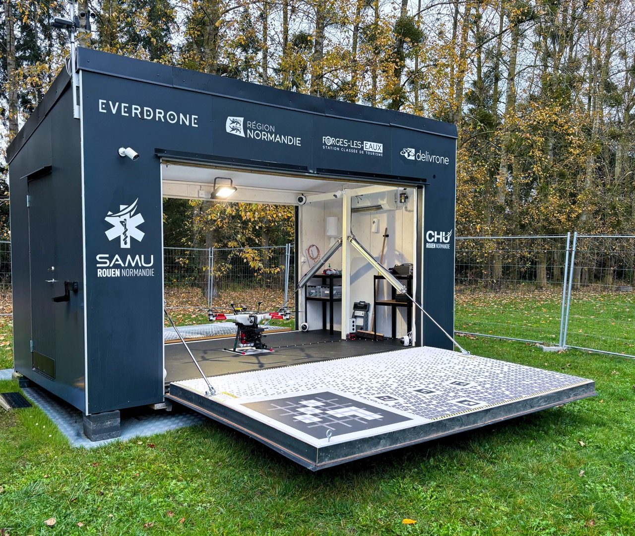 A black Everdrone emergency drone hangar sits on a grassy field with autumn trees in the background. The front of the hangar is fully open, revealing a drone positioned on its landing platform, which extends outward like a ramp. The structure is branded with logos including Everdrone, Région Normandie, Forges-les-Eaux, Delivrone, SAMU Rouen Normandie, and CHU Rouen Normandie. Inside the hangar, equipment shelves and lighting are visible. The area is enclosed by fencing.